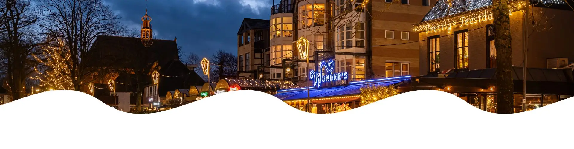 Luchtfoto van Bergen aan Zee met een uitgestrekt duinlandschap, een dorp met huizen en gebouwen, en een breed zandstrand langs de kustlijn. De zon weerspiegelt op de zee onder een blauwe lucht met wolken.