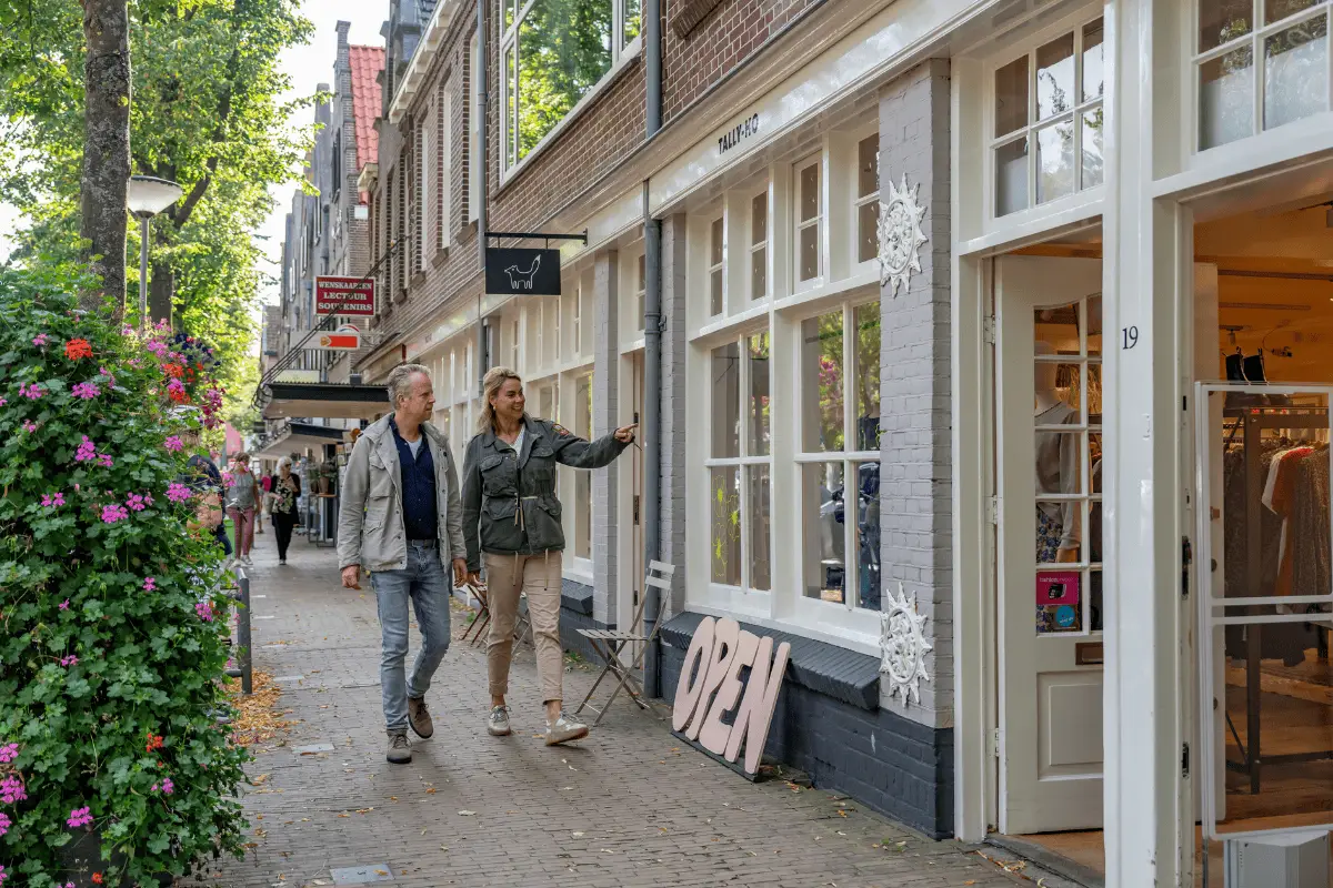 Twee mensen lopen langs boetieks en winkels in een sfeervolle winkelstraat met bomen en historische gevels.