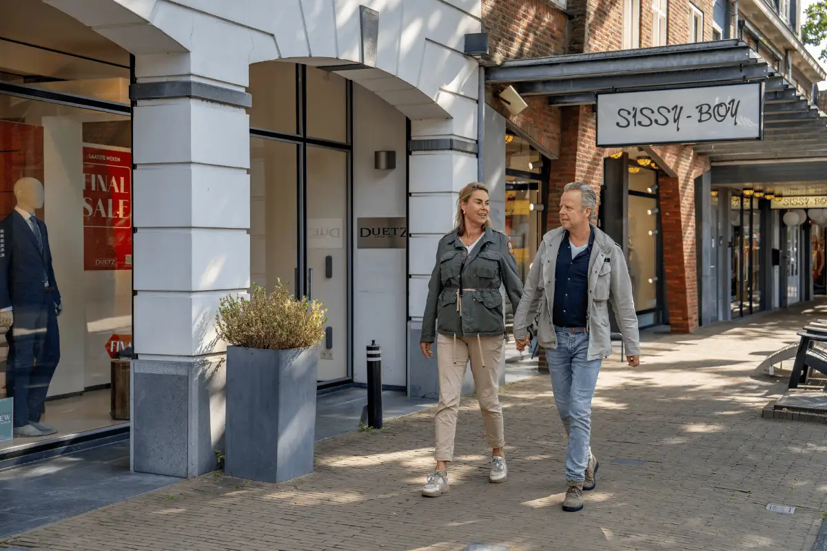 Twee mensen wandelen langs winkels in een winkelstraat met etalages en boetieks.