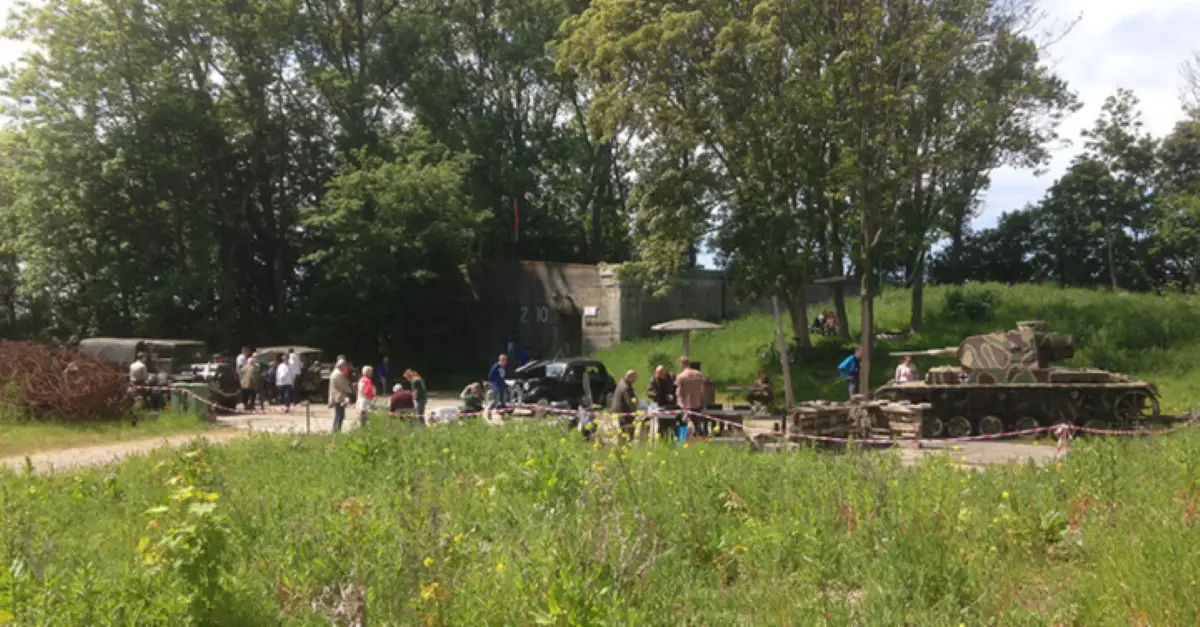 Buitenlocatie bij Museum Vliegveld Bergen met een bunker op de achtergrond en historische militaire voertuigen, waaronder een gecamoufleerde tank. Bezoekers lopen rond en bekijken de tentoonstellingen in een groene, bosrijke omgeving.