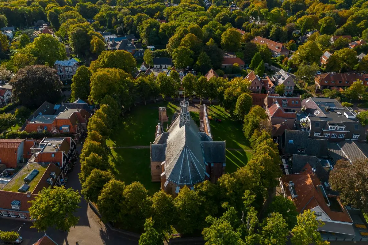 Luchtfoto van de Ruïnekerk in Bergen, omgeven door groen en een sfeervol dorp met huizen en winkels. De kerk en ruïnes liggen centraal tussen de bomen, onder een helderblauwe lucht met enkele wolken.