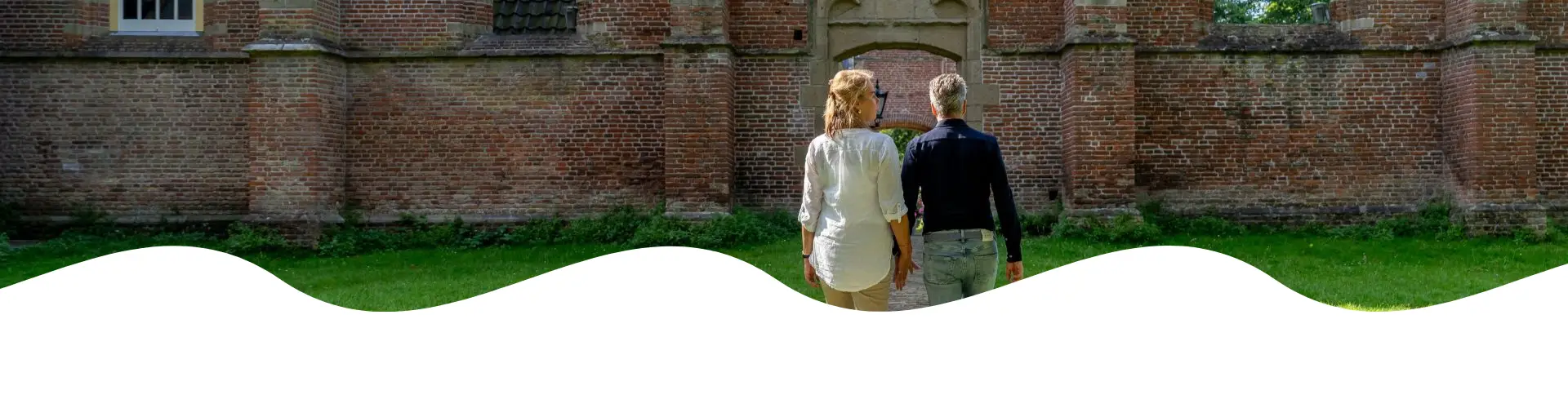 Een rustig bospad omgeven door hoge, groene bomen met zonlicht dat door het bladerdak schijnt. Twee mensen wandelen samen over het met bladeren bedekte pad in een serene, natuurlijke omgeving.