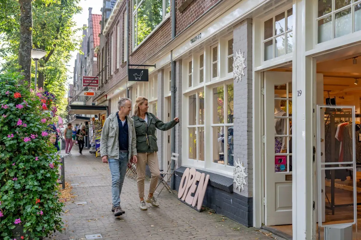 Een levendige winkelstraat met charmante bakstenen panden en grote ramen. Mensen wandelen en winkelen, terwijl fietsen geparkeerd staan langs de stoep. In het midden loopt een stel hand in hand over een zebrapad.