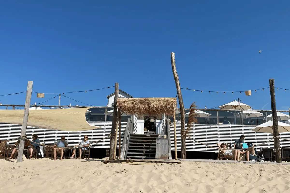 Strandpaviljoen op het zand met een rieten entree, terrastafels met bezoekers en een heldere blauwe lucht boven zee en strand.