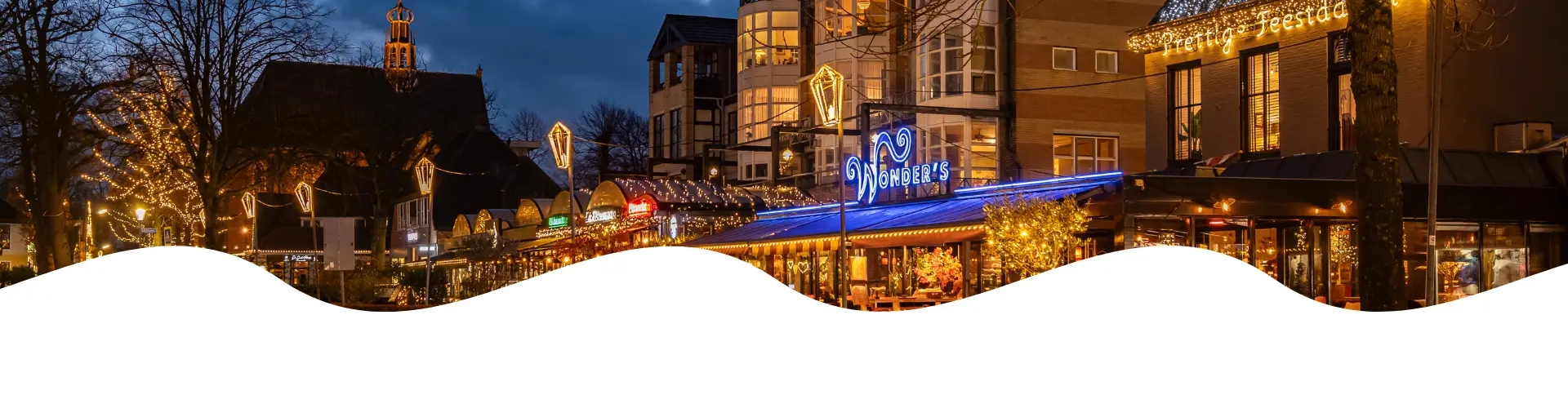 Gezellige straat in de avond met verlichte restaurants en cafés, kerstverlichting in de bomen en nat wegdek dat het warme licht van de gebouwen weerspiegelt.