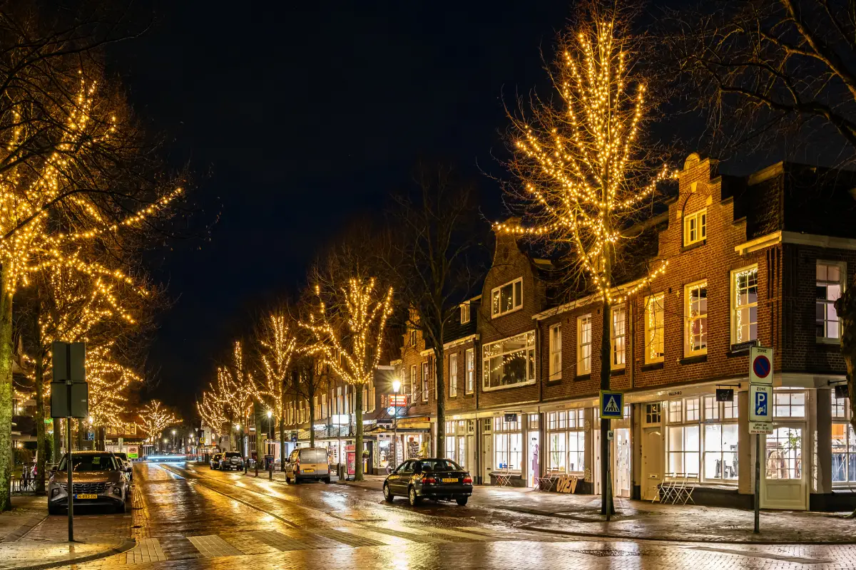 Sfeervol verlichte winkelstraat in de avond met kerstverlichting in de bomen, nat wegdek dat licht reflecteert en verlichte etalages in historische panden.