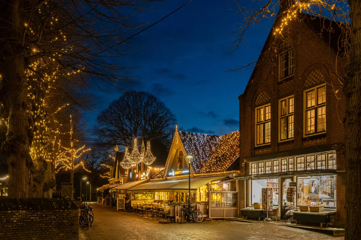 Sfeervol verlicht dorpscentrum in de avond met restaurants en winkels onder warme kerstverlichting, met historische gebouwen en een blauwe avondlucht.