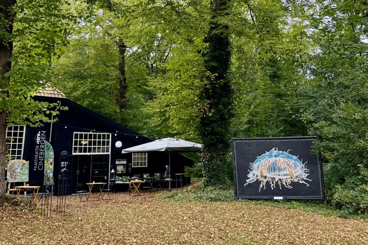 Paviljoen van Filmtheater Cinebergen in een bosrijke omgeving met terras en een groot kunstwerk langs het pad.