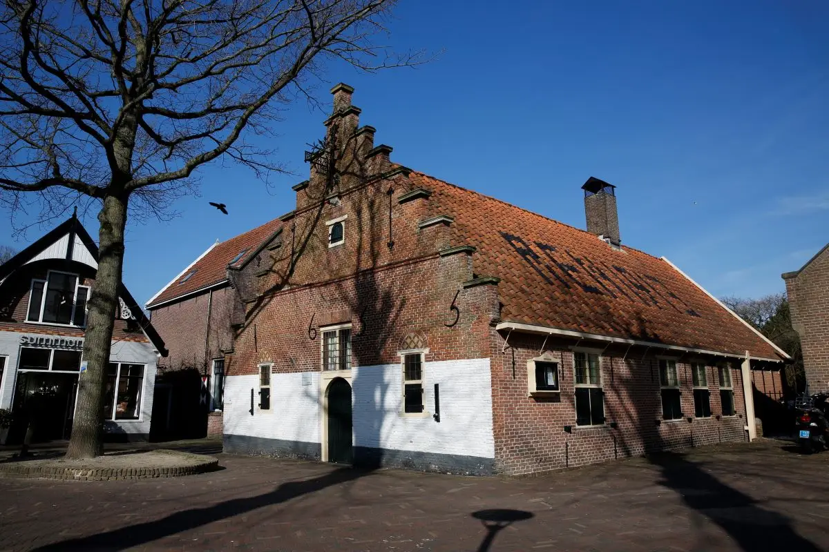 Historisch pand met trapgevel in Bergen. Het gebouw heeft een rood pannendak en deels witgekalkte bakstenen muren, met jaartallen en een inscriptie op het dak. Op de voorgrond een geplaveide straat en een boom met kale takken onder een blauwe lucht.