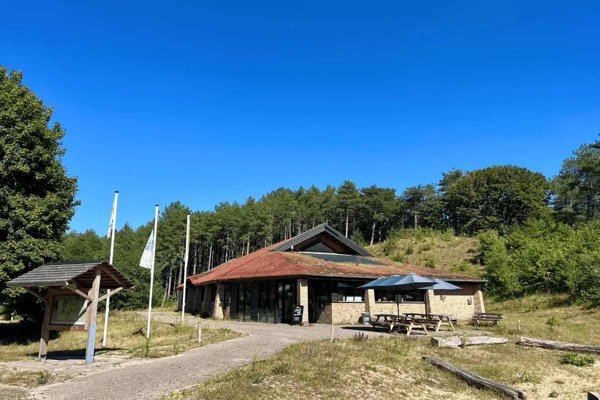 Bezoekerscentrum in een duingebied met een gebouw, picknicktafels en bos op de achtergrond onder een heldere blauwe lucht.