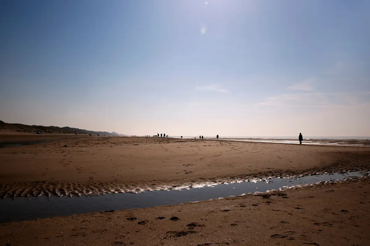 Een uitgestrekt strand in Bergen aan Zee met een smalle watergeul in het zand. Op de achtergrond wandelen silhouetten van mensen langs de kustlijn onder een heldere, blauwe lucht.