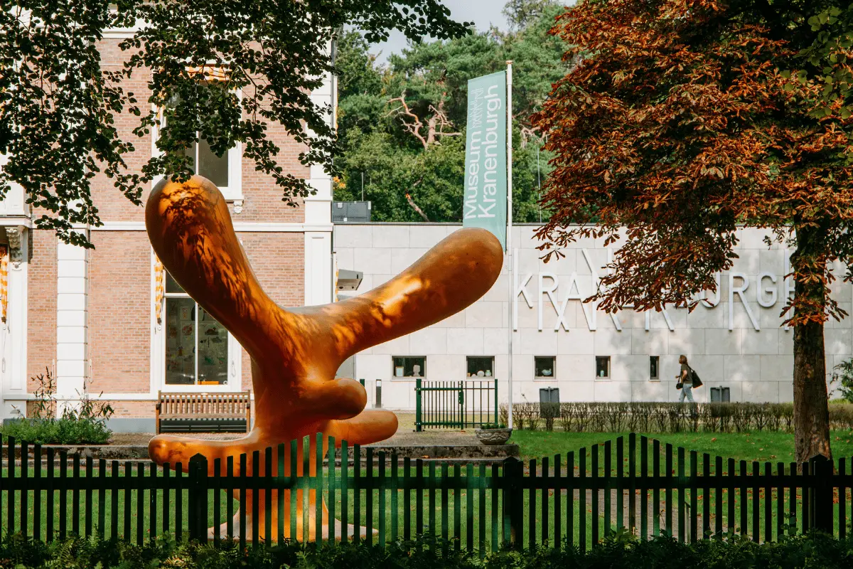 Groot oranje kunstwerk in een park voor cultureel centrum De Kranenburgh, met bomen, gras en het gebouw op de achtergrond.