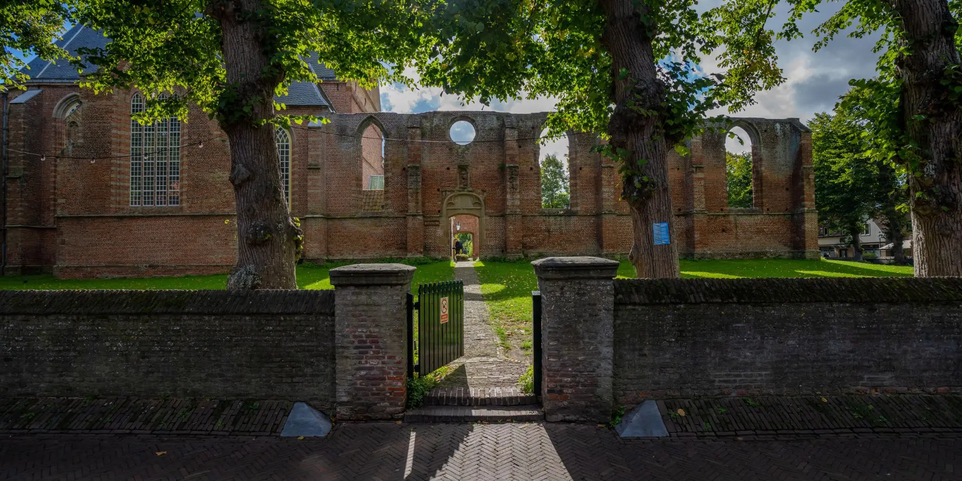 Ingang van de Ruïnekerk in Bergen, met een open poort en een pad dat leidt naar de overgebleven bakstenen muren met grote gotische ramen. De zon schijnt door de groene bomen, waardoor schaduwen op de bestrating en het grasveld vallen.