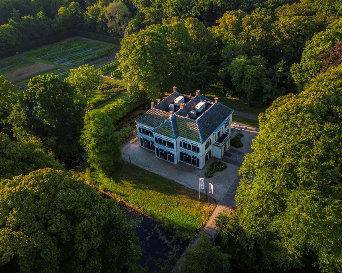 Luchtfoto van Het Hof in Bergen, een statig landhuis met een blauw pannendak, omringd door een groene tuin en een bosrijke omgeving. Voor het huis ligt een oprijlaan met vlaggen, en in de achtergrond zijn moestuinen en aangelegde paden zichtbaar.