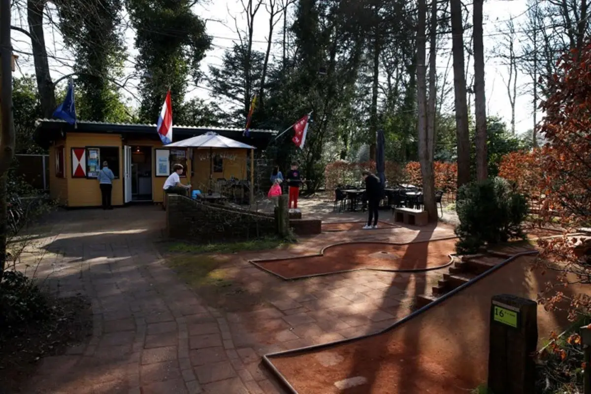 Midgetgolfbaan Merelhof, gelegen in een bosrijke omgeving met hoge bomen. Op de voorgrond een midgetgolfbaan met rode banen en hindernissen, terwijl bezoekers spelen. Op de achtergrond een geel houten gebouw, waar mensen zich aanmelden.
