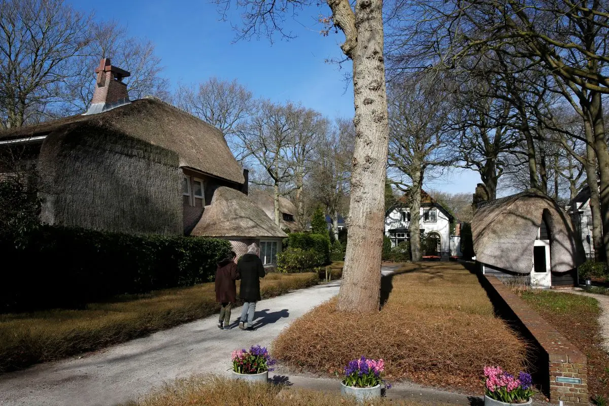 "Park Meerwijk met een idyllische laan omgeven door rietgedekte villa’s in Amsterdamse School-stijl. Twee mensen wandelen over een grindpad, omringd door bomen en bloeiende voorjaarsbloemen.