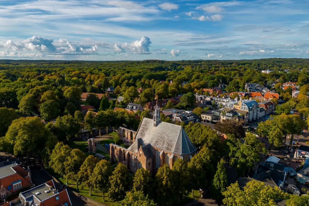 Luchtfoto van de Ruïnekerk in Bergen, omgeven door groen en een sfeervol dorp met huizen en winkels. De kerk en ruïnes liggen centraal tussen de bomen, onder een helderblauwe lucht met enkele wolken.