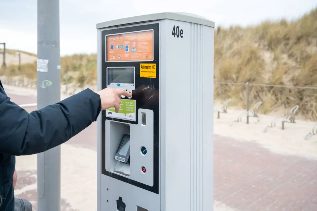 Een parkeerautomaat in Bergen aan Zee met een persoon dat aan het betalen is op het automaat, met op de achtergrond het duin en fietsenstallingen