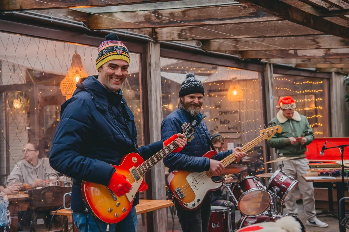 Muzikanten spelen buiten onder een overkapping tijdens een winterse setting. De gitarist en bassist dragen warme kleding en mutsen, terwijl er op de achtergrond een drummer en een toetsenist te zien zijn. De sfeer is informeel en gezellig met lichtslingers en houten details.