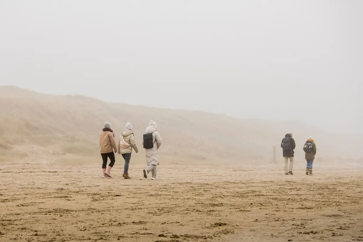 Groep mensen wandelt over een breed, verlaten strand op een mistige dag, met duinen op de achtergrond en gekleed in warme winterkleding.