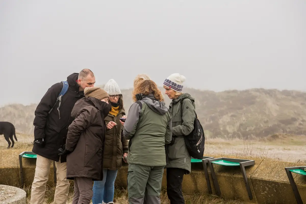 Groep wandelaars in winterkleding staat samen rond een informatiepunt in de mistige duinen, verdiept in een kaart of uitleg, met heuvelachtig duinlandschap op de achtergrond.