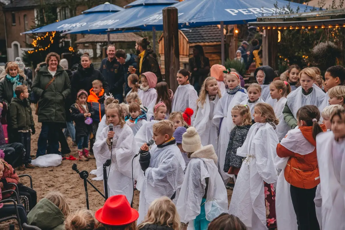 Groep kinderen zingt buiten in witte capes voor publiek, onder grote parasols en met toekijkende ouders in winterkleding.