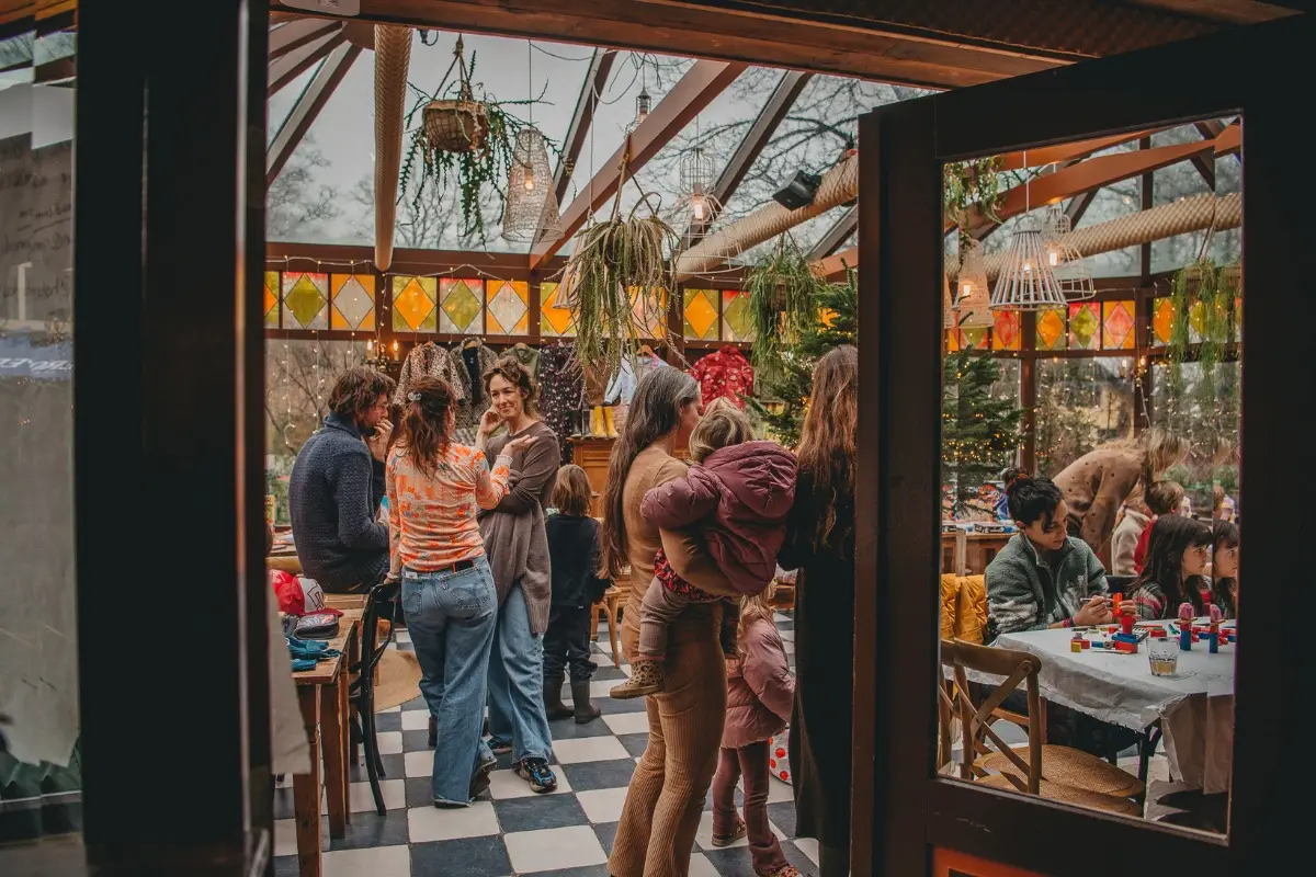 Gezellige drukte in een knusse, met planten versierde serre waar volwassenen en kinderen samenkomen voor een winterse bijeenkomst. Aan tafels wordt geknutseld, terwijl anderen staand praten en kinderen spelen. De sfeer is warm en feestelijk met kleurrijke raampanelen, kerstversiering en zachte verlichting.