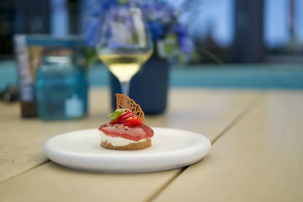 Fijn dessert met aardbei en decoratief suikerwerk op een wit bord, geserveerd op een houten tafel met op de achtergrond een glas witte wijn en blauwe bloemen in vaasjes.