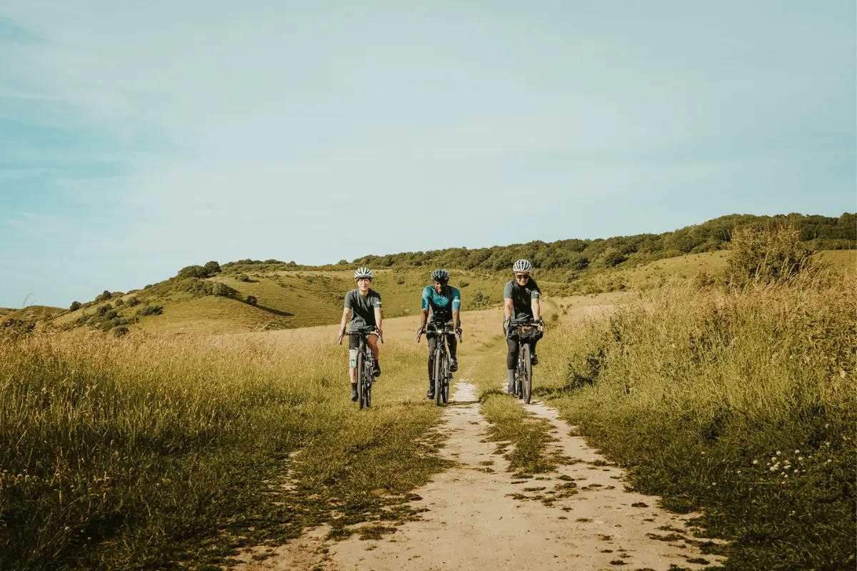 Drie fietsers rijden over een zandpad door een heuvelachtig duinlandschap met gras en struiken onder een heldere lucht.
