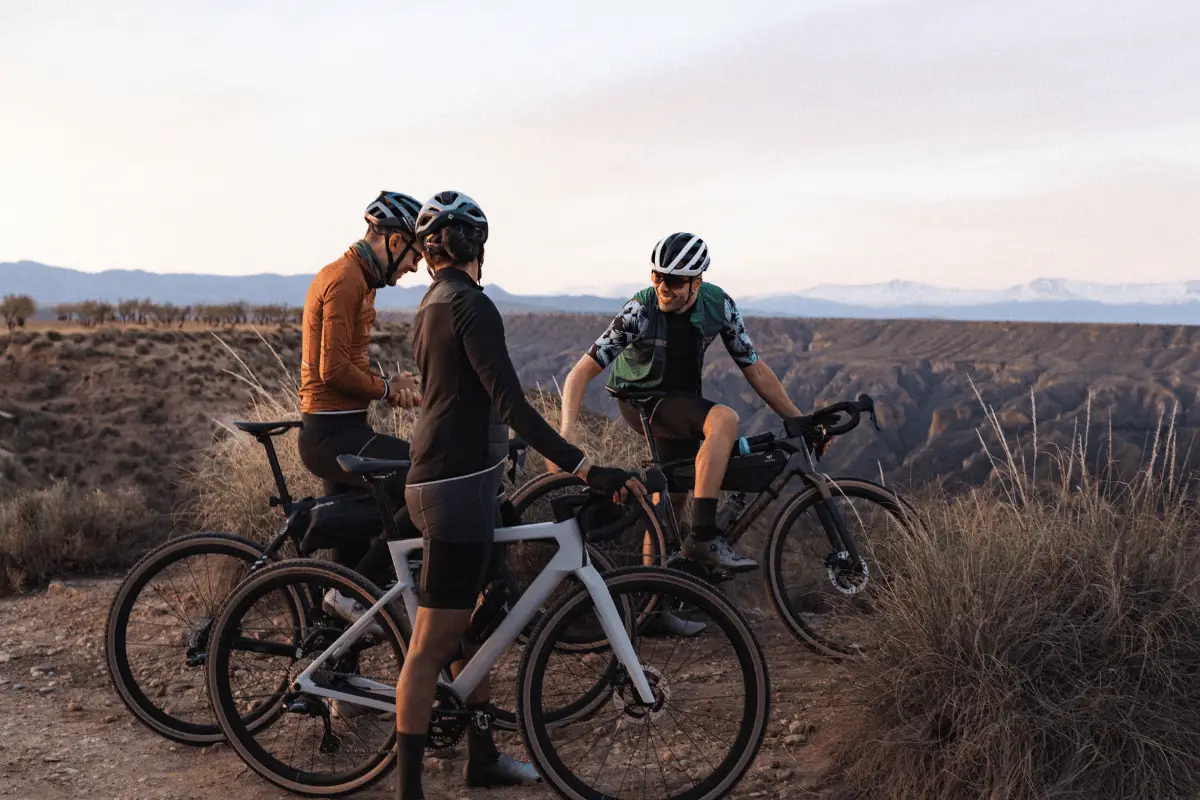 Drie wielrenners staan met hun fietsen op een uitkijkpunt in een heuvelachtig landschap en praten samen terwijl ze van het uitzicht genieten.