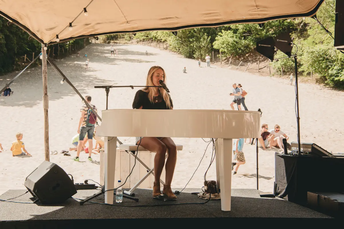 Zangeres speelt piano en zingt op een podium aan de voet van een zandduin, terwijl bezoekers op de achtergrond genieten van het optreden.