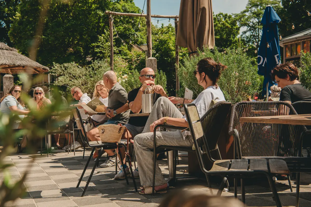 Mensen ontspannen op een zonnig terras tussen het groen, met tafels, stoelen en parasols in een sfeervolle buitenomgeving.