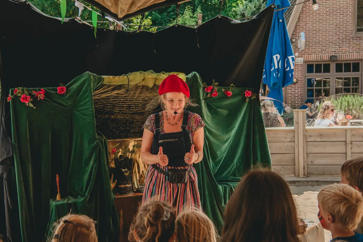 Vertelster geeft een voorstelling aan kinderen in een kleine openluchtsetting met decor en publiek op de voorgrond.