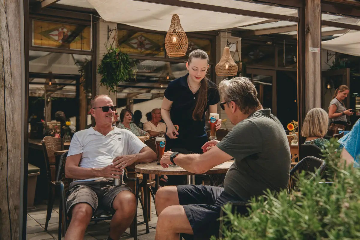 Bediening serveert drankjes aan gasten op een gezellig terras met tafels, planten en warme verlichting.