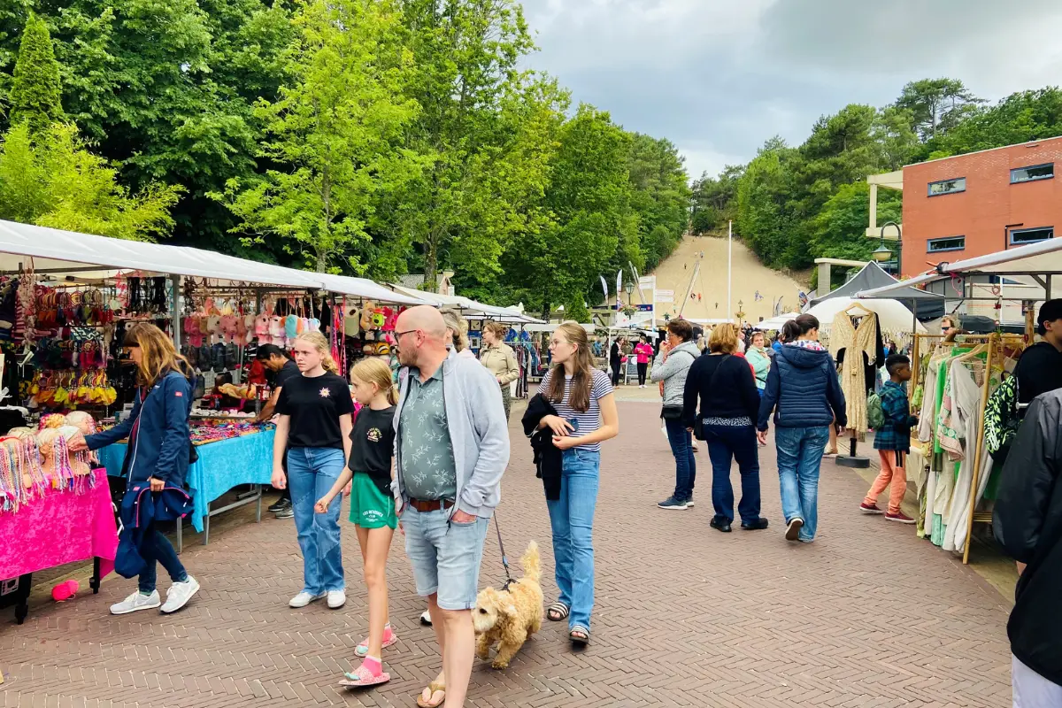 Drukke markt in een Schoorl met kraampjes aan weerszijden van de straat, bezoekers die langs de stands lopen en winkelen, met op de achtergrond een hoge duin met trap.