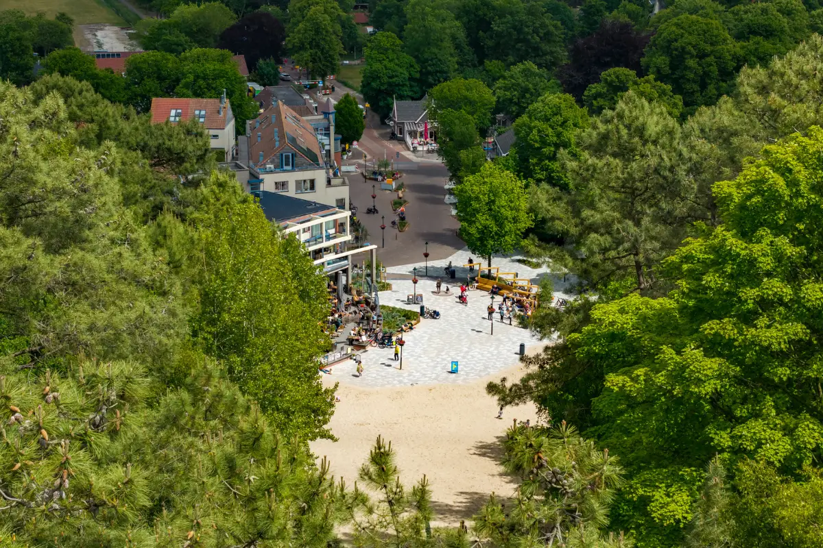 Uitzicht vanaf een duintop in Schoorl op een plein met terras en bezoekers, omringd door groene bomen en huizen. Een zandpad tussen de bomen leidt vanaf de duinen naar het dorpscentrum.