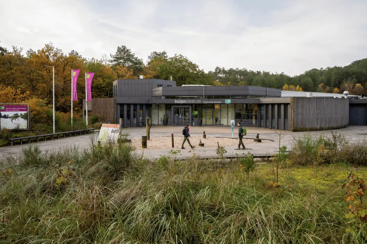 Bezoekerscentrum Schoorlse Duinen met ingang, vlaggen en wandelaars ervoor, omringd door duinvegetatie en bos.