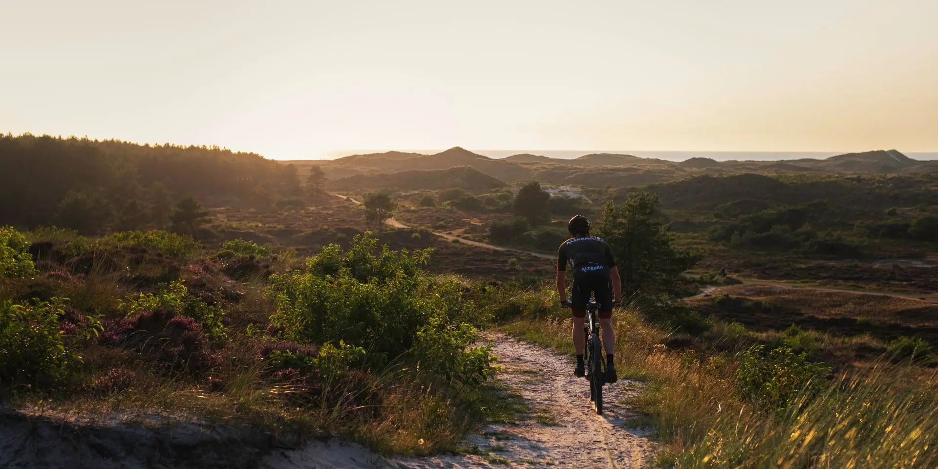 Een mountainbiker fietst over een zandpad door een heuvelachtig duinlandschap bij zonsondergang. De warme gloed van de ondergaande zon verlicht de heide en struiken, met in de verte glooiende duinen en een heldere horizon.