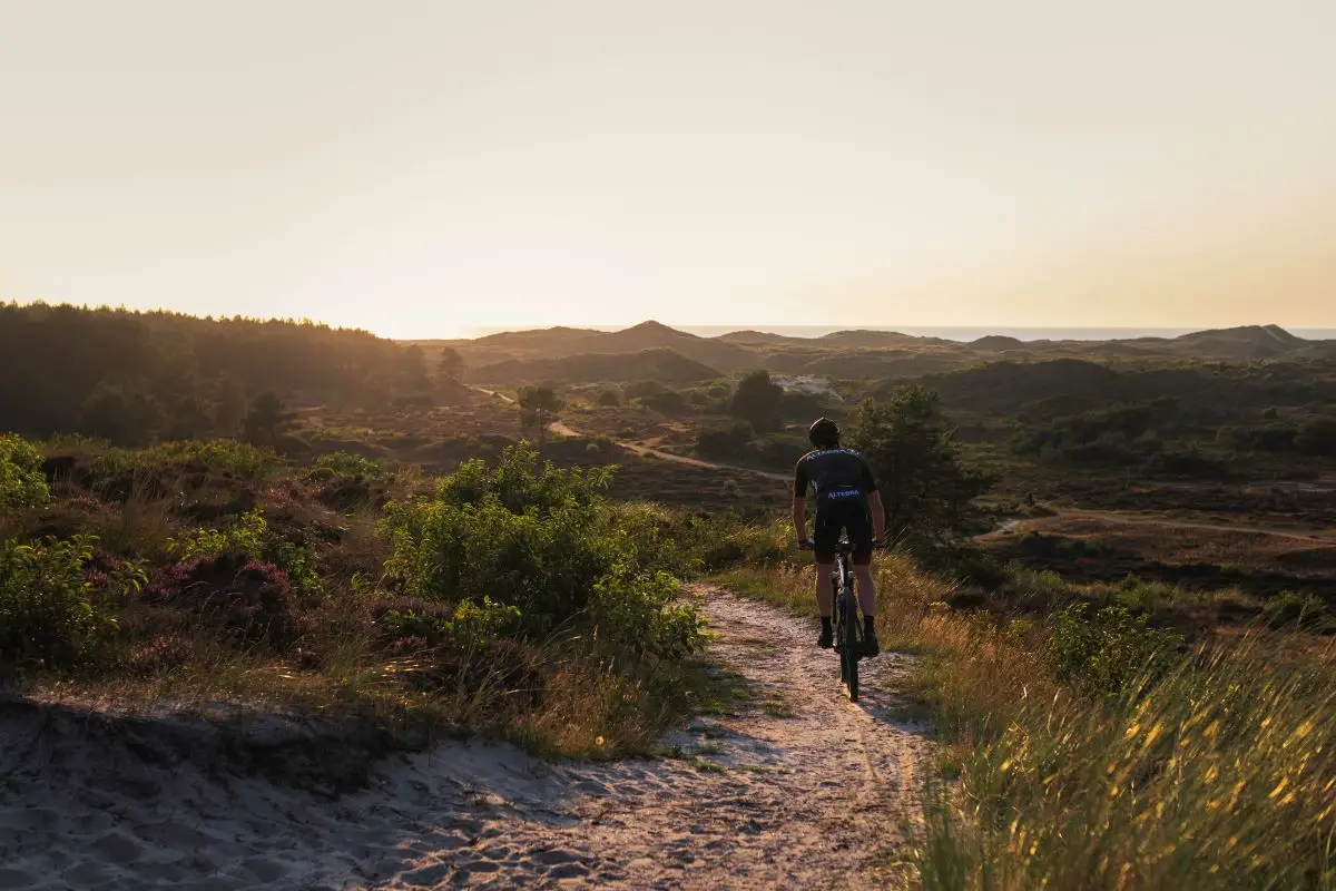 Een mountainbiker fietst over een zandpad door een heuvelachtig duinlandschap bij zonsondergang. De warme gloed van de ondergaande zon verlicht de heide en struiken, met in de verte glooiende duinen en een heldere horizon.