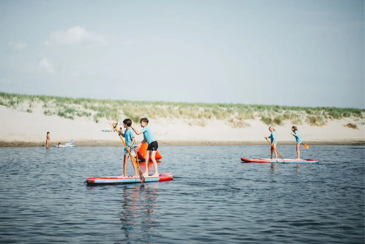 Kinderen peddelen op rode paddleboards in de lagune van Camperduin. Op de achtergrond liggen zandduinen en een paar zwemmers genieten van het water onder een lichte, zomerse lucht.