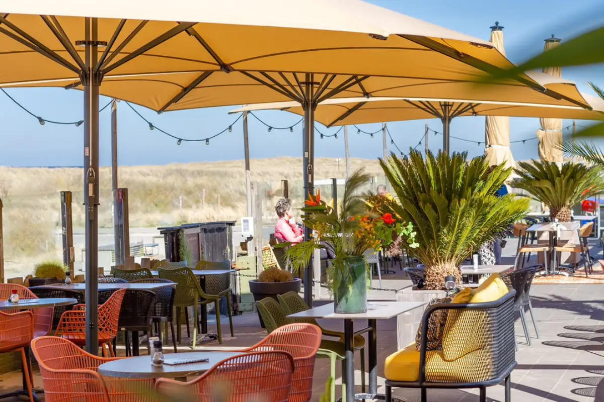 Het buiten terras bij strandpaviljoen Struin met gekleurde stoelen, kussens, parasols, gezellige lichtjes en tafels. Met uitzicht op het duingebied.