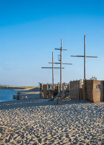 Houten speeltoestel in de vorm van een schip op het strand, met zand, duinen en water onder een heldere blauwe lucht.