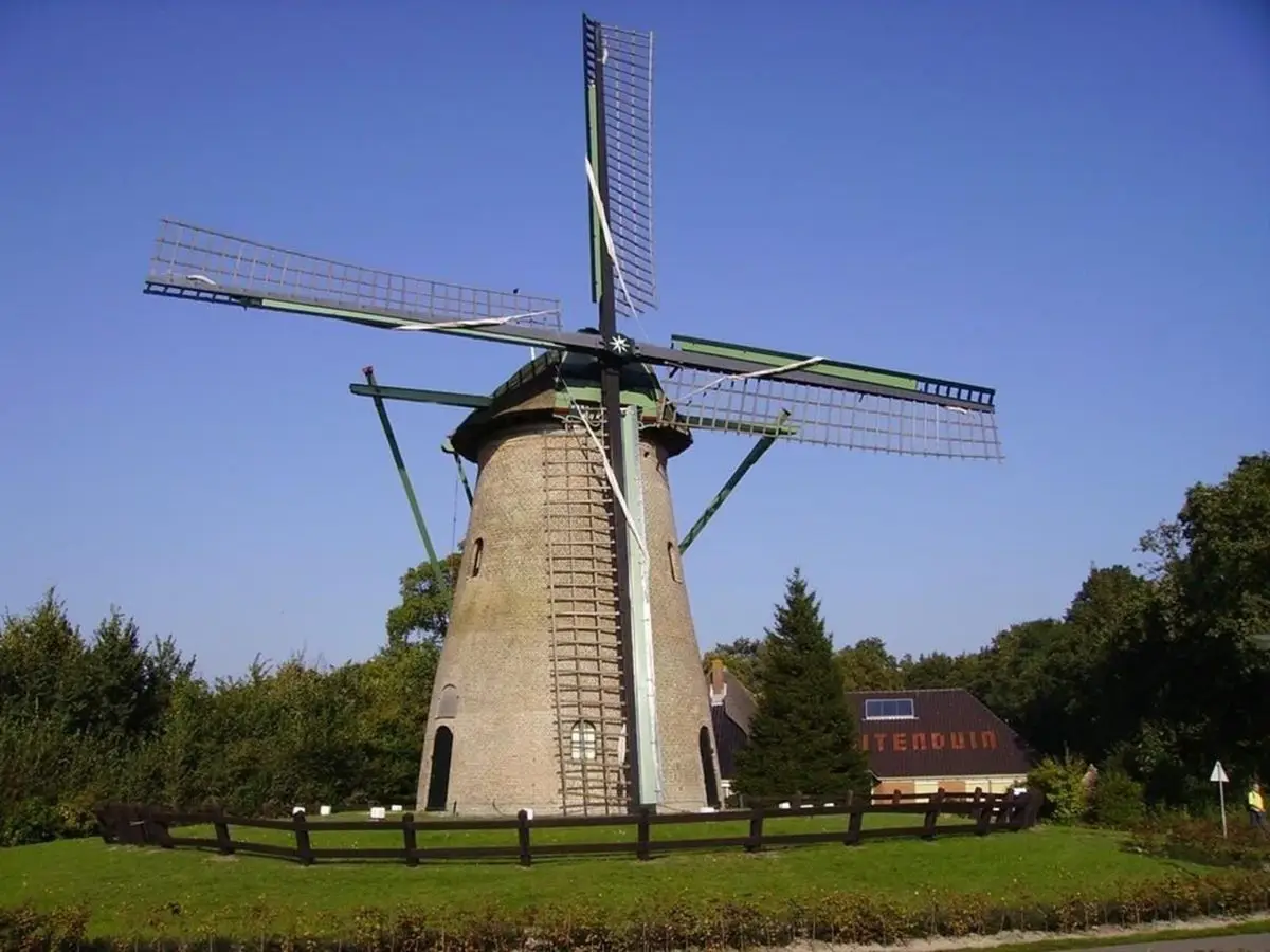 Molen Kijkduin, een traditionele Nederlandse windmolen met een stenen romp en groene wieken, omgeven door een laag houten hek en groen gras. Op de achtergrond staat een gebouw met de naam 'Kijkduin' in een bosrijke omgeving onder een helderblauwe lucht