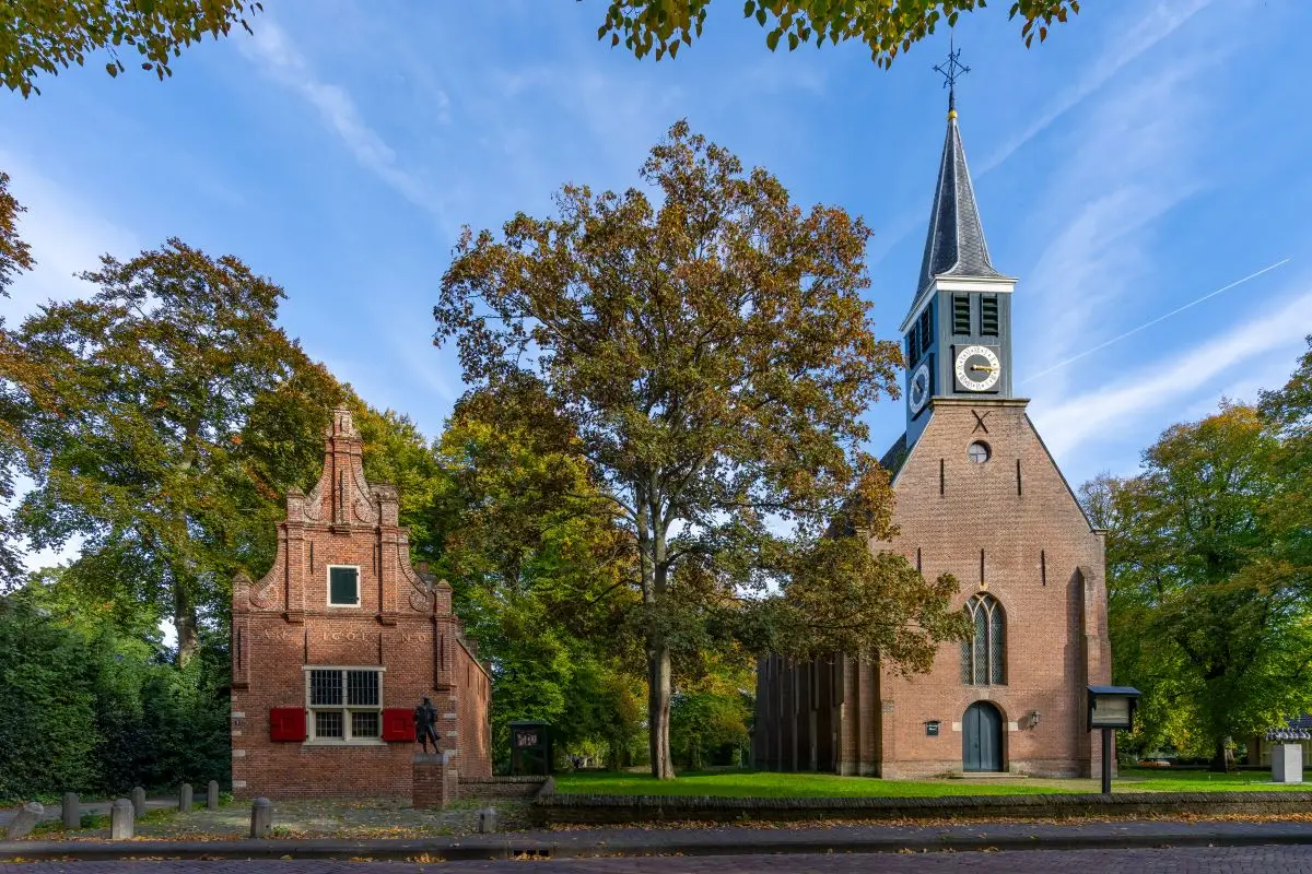 De kerk en het raadhuis van Schoorl, omringd door groene bomen. De kerk heeft een bakstenen gevel met een hoge spits en een klok, terwijl het raadhuis een trapgevel en rode luiken heeft. Een beeld van een figuur staat bij de ingang van het raadhuis