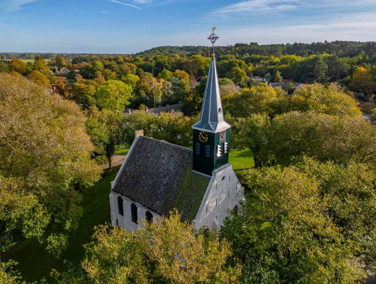 Bovenaanzicht van het witte kerkje, een wit geschilderd, karakteristiek kerkje met groene accenten, tussen de herfst getinte bossen in Schoorl op een zomerse dag