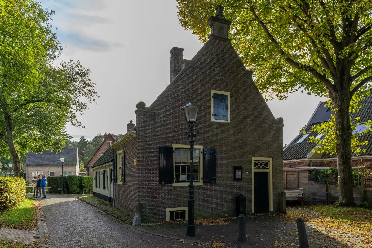 Het historische raadhuis van Groet, een karakteristiek bakstenen gebouw met een trapgevel en zwarte luiken. Een oude lantaarnpaal staat voor het pand, en de straat wordt omringd door bomen. In de achtergrond wandelen mensen door het dorp.