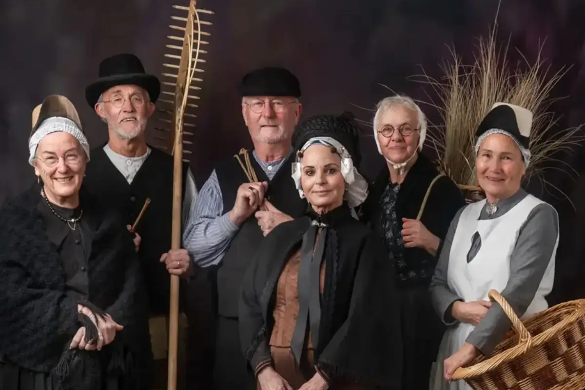 Groep Egmondse derpers in traditionele klederdracht poseert samen voor een donkere achtergrond. De mannen en vrouwen dragen authentieke kleding en gebruiken zoals een hark, mand en bundel duingras, waarmee de rijke historie en verbondenheid van Egmond zichtbaar worden.
