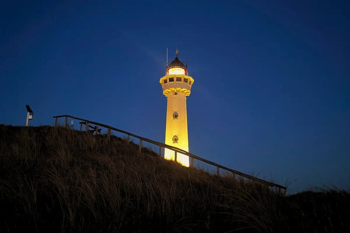 De verlichte vuurtoren J.C.J. van Speijk torent boven de duinen van Egmond aan Zee uit tijdens de schemering. Het warme licht van de iconische vuurtoren steekt af tegen de diepblauwe avondlucht en benadrukt de rustige, karaktervolle sfeer van de kustplaats.