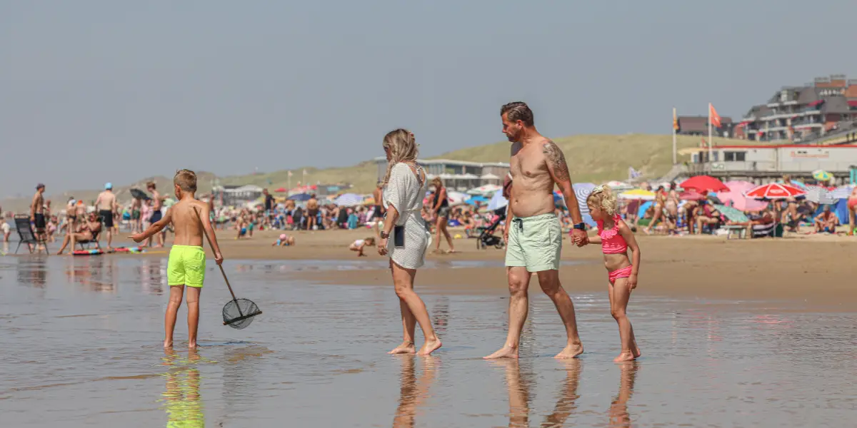 Mensen wandelen over een duinpad richting strand in Egmond aan Zee op een zonnige dag. Links staan strandpaviljoens en rieten lampenkappen hangen aan palen langs het pad. Op de achtergrond is de zee zichtbaar met windmolens aan de horizon.