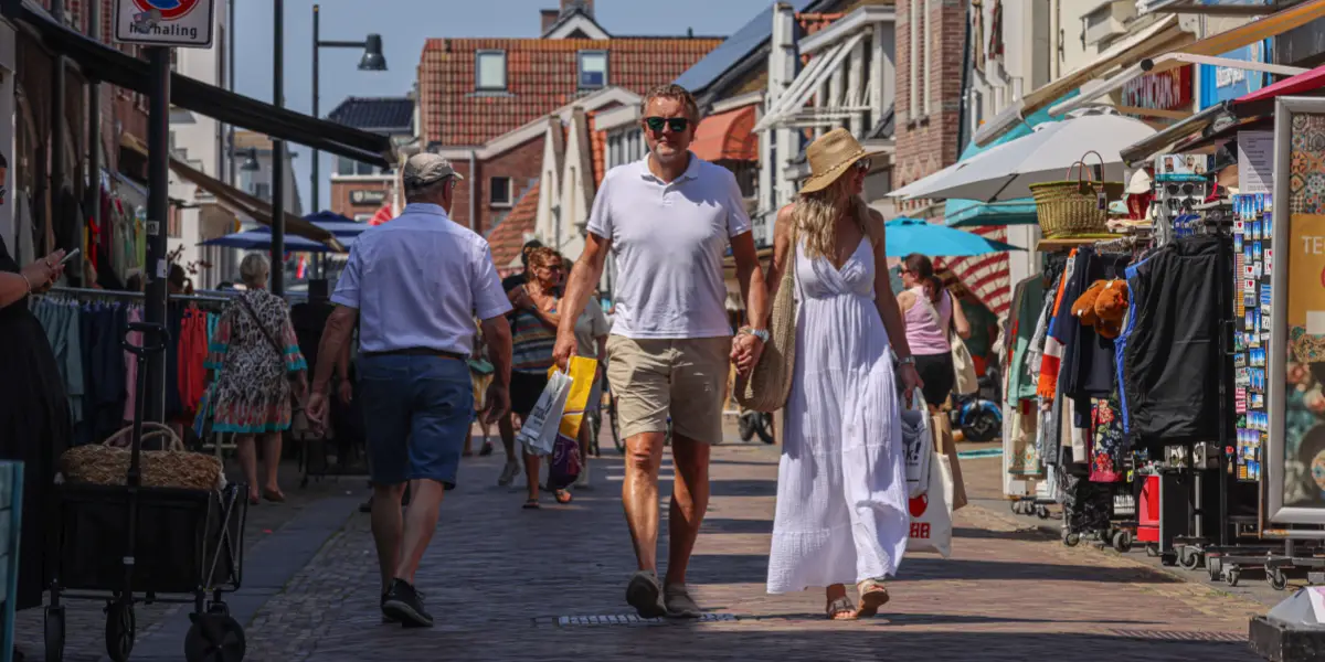Zonnige winkelstraat aan zee in Egmond met wandelaars en toeristen op een geplaveid plein. Op de voorgrond staan kleurrijke tulpen in een metalen ton. Op de achtergrond staan kraampjes met souvenirs en kleurrijke ballonnen, met winkels en terrassen onder een blauwe lucht.